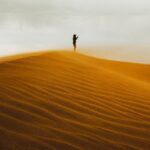 A lone figure stands on a sand dune at sunset.