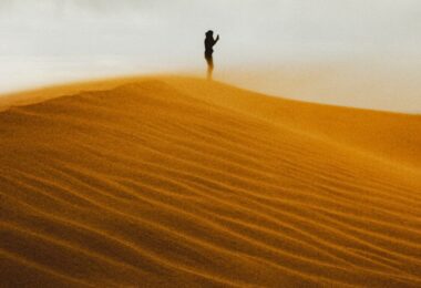 A lone figure stands on a sand dune at sunset.